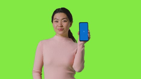 studio portrait of smiling woman holding blue screen mobile phone towards camera against green screen 3