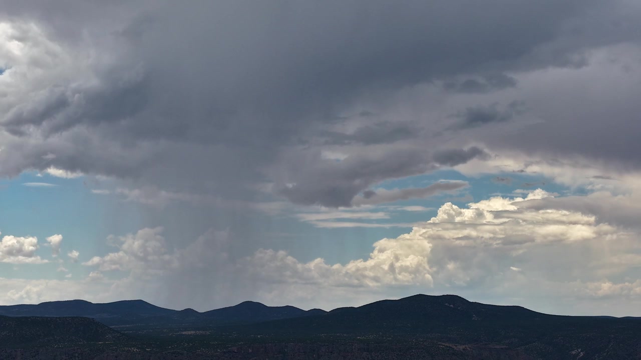 4K drone timelapse providing a closer view of a localized downpour. A distinct rain shaft falls from a heavy grey cloud canopy onto dark, rolling mountains, contrasted by bright white background cloud