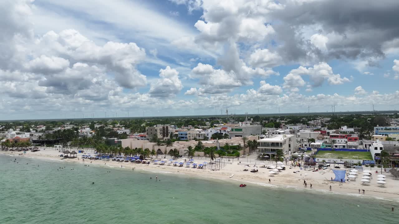 A lateral drone flight over Puerto Progreso, showing the beachfront city with crowded sandy shores, turquoise water, and dramatic towering clouds on a bright sunny day