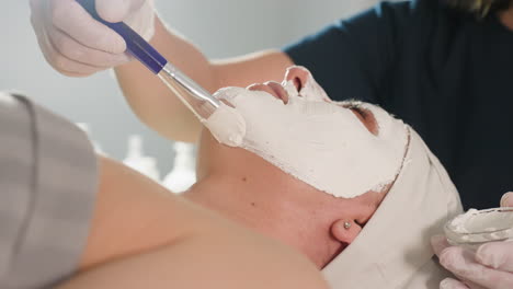 cosmetologist smooths in circular motions thick face mask carefully with soft silicone brush on face of spa visitor under professional bright light during relaxing skin treatment session