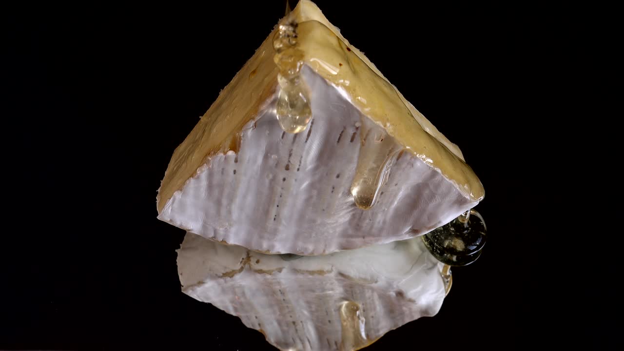 капли меда, капающие с сыра бри на зеркальном черном фоне.