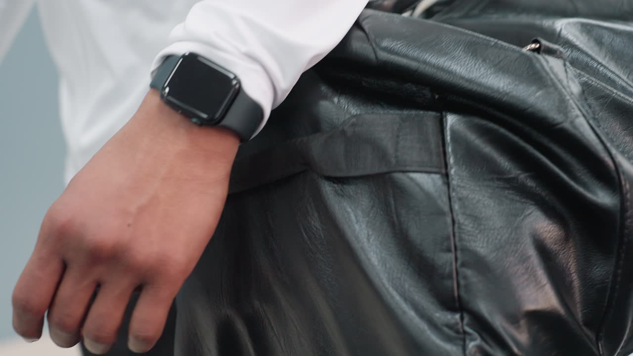 close up of man wearing black smartwatch and white athletic top carrying large leather duffel bag and holding wireless headphones while walking through bright modern gym corridor with equipment