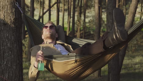 a relaxed guy takes a drink lying in a hammock in the middle of the forest