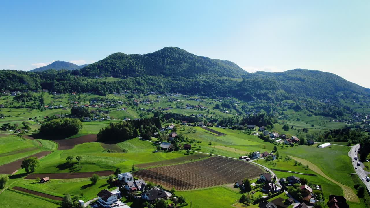 Aerial Over Rolling Green Fields And Hills With Forested Mountain In The Background