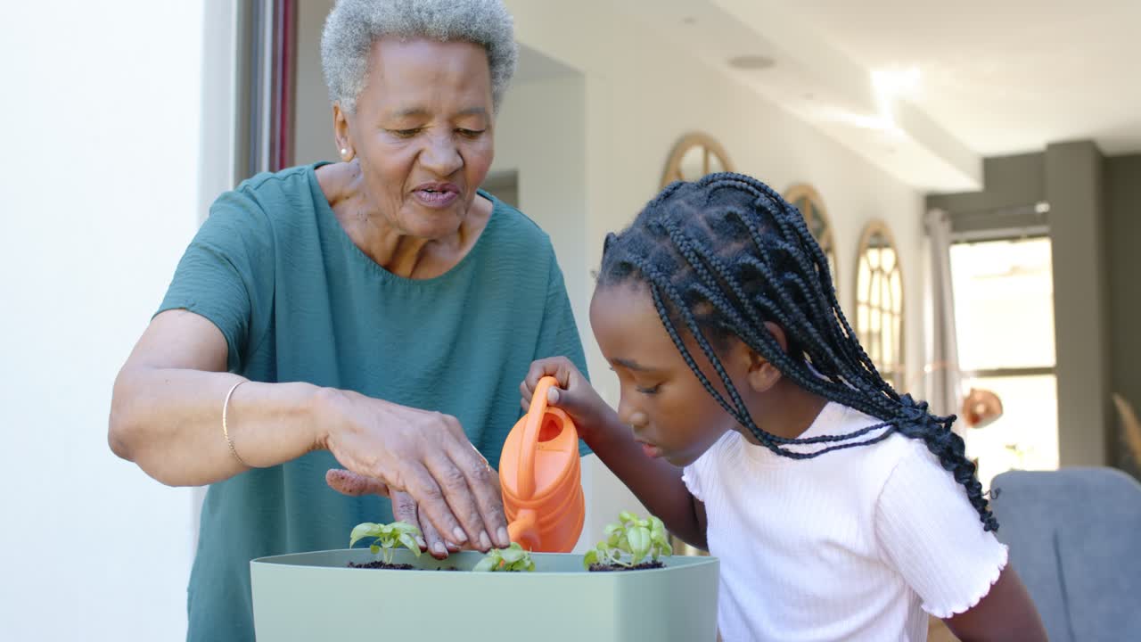 Senior African woman teaching youth girl at porch planter, girl pouring from orange watering can