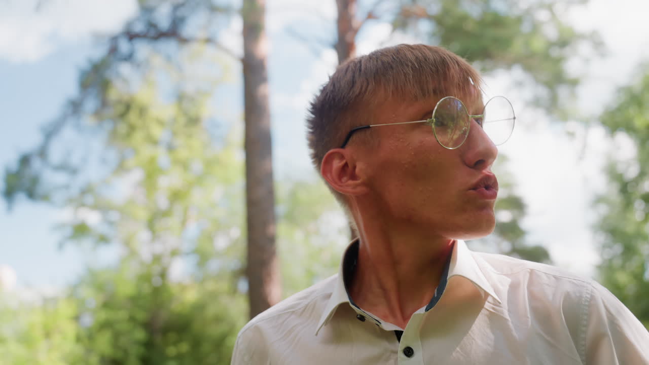 Portrait view of man in white shirt and glasses smiling warmly while walking through forest path, observing tall trees and enjoying bright atmosphere of summer day surrounded by greenery and sunlight