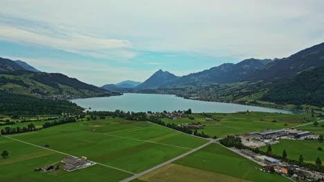 Vista-Aérea-De-Un-Remoto-Pueblo-Frente-Al-Lago,-Rodeado-De-Exuberantes-Campos-Verdes-Y-Montañas,-Con-Una-Cima-Alta-En-La-Distancia