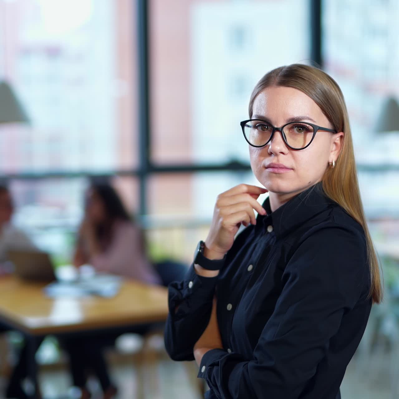 Young female employee standing in office thinking over something. Side view. Two people talking at the table in blur at the background