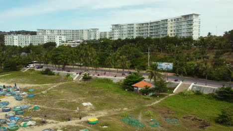 Aerial-view-of-scooter-driving-along-coastline-highway-with-residential-hotel-district-carrying-long-surf-board-for-practicing-in-ocean-sealinks,-vietnam