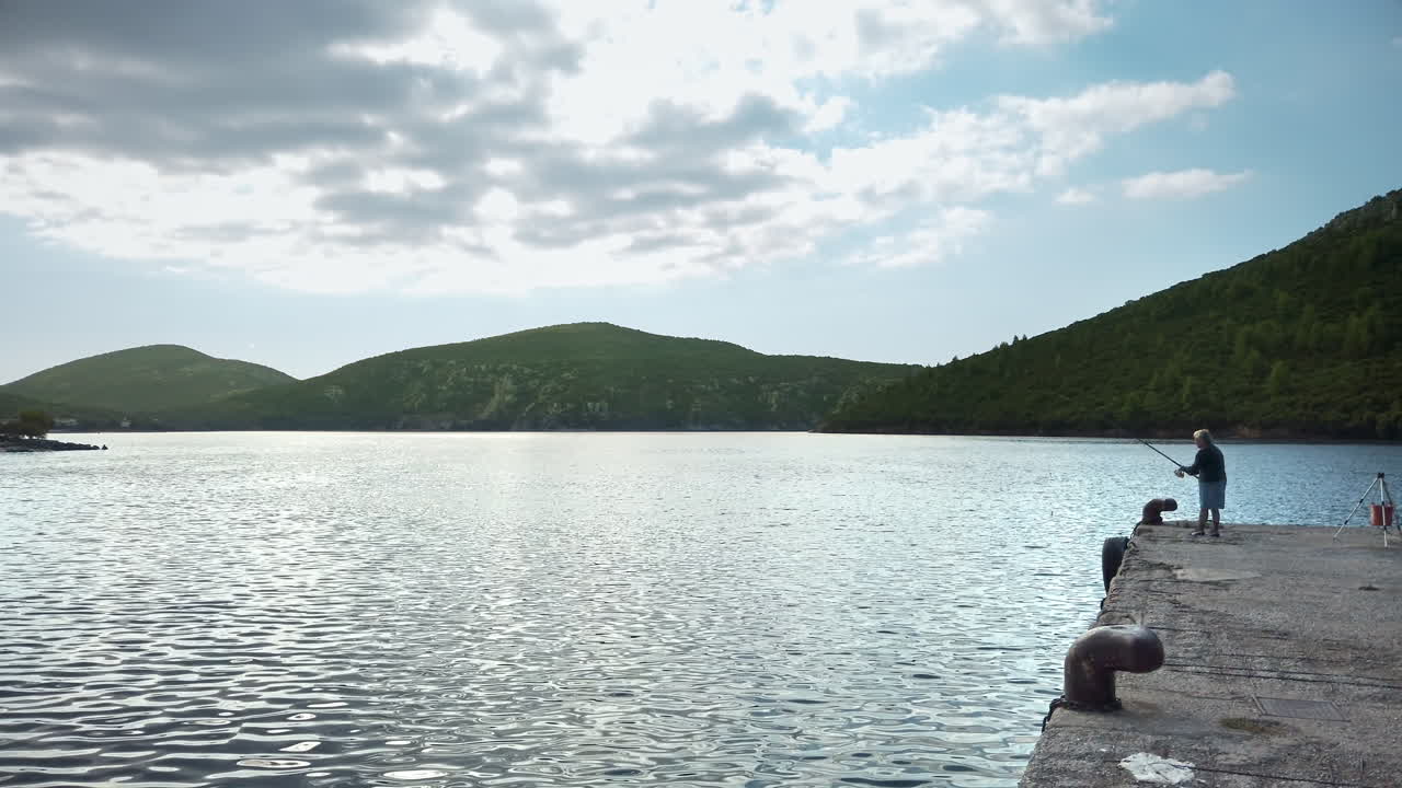 Fishing man on the pier in a village, a lot of greenery, green hills on the background, Greece. Slow motion