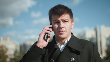 freelancer in black coat engaged in phone call outdoors, shaking head in disapproval while reacting to discussion, background features clear blue sky, modern architecture, and blurred urban greenery