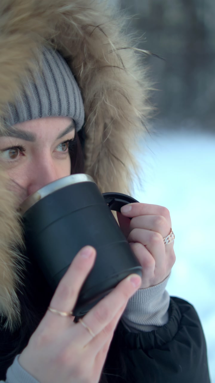 A woman stands in the snow holding a black mug. She takes a moment to savor the warm drink. The scene captures the cold winter setting with snow all around her.