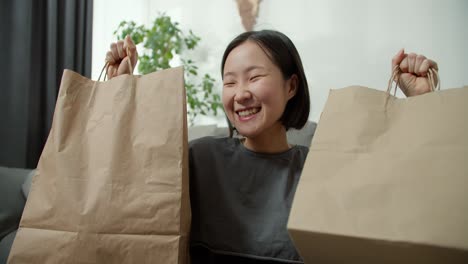 Señora-Asiática-Emocionada-Comprando-En-Línea,-Levantando-Bolsas-De-Regalo-Sobre-Su-Cabeza-En-Casa