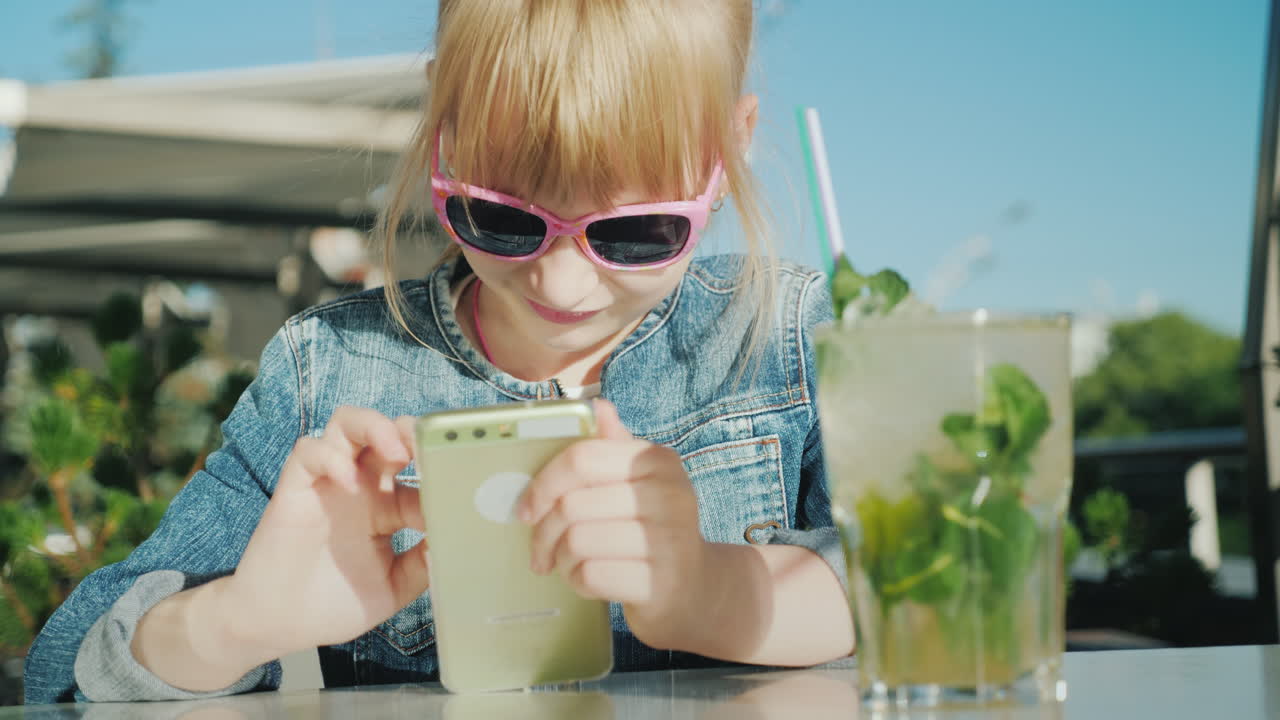 분홍색 안경을 쓴 우스꽝스러운 소녀가 스마트폰으로 노는 것은 카페의 여름 아파트에 앉아 있다