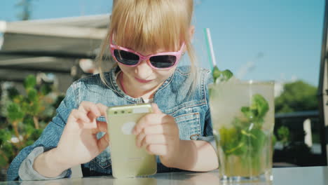 Lustiges-Mädchen-In-Rosa-Brille-Spielt-Auf-Dem-Smartphone-Sitzt-Auf-Einer-Sommerwohnung-In-Einem-Café