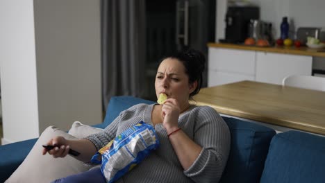 Overweight-woman-sitting-on-couch-at-home,-watching-tv-and-eating-potato-chips