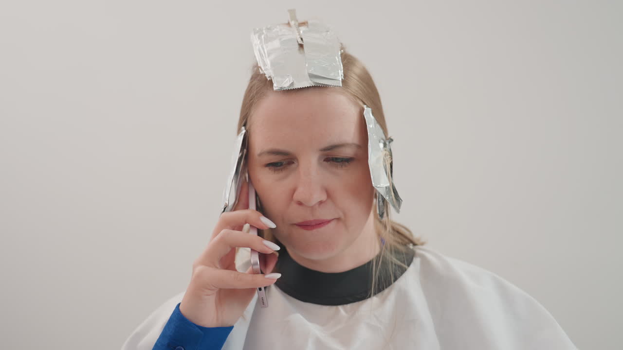 Mujer caucásica en una peluquería con papel de aluminio, hablando por teléfono mientras le hacen mechas, con una capa blanca, fondo neutro, primer plano, expresión concentrada, secciones de papel de aluminio en el pelo, estilista