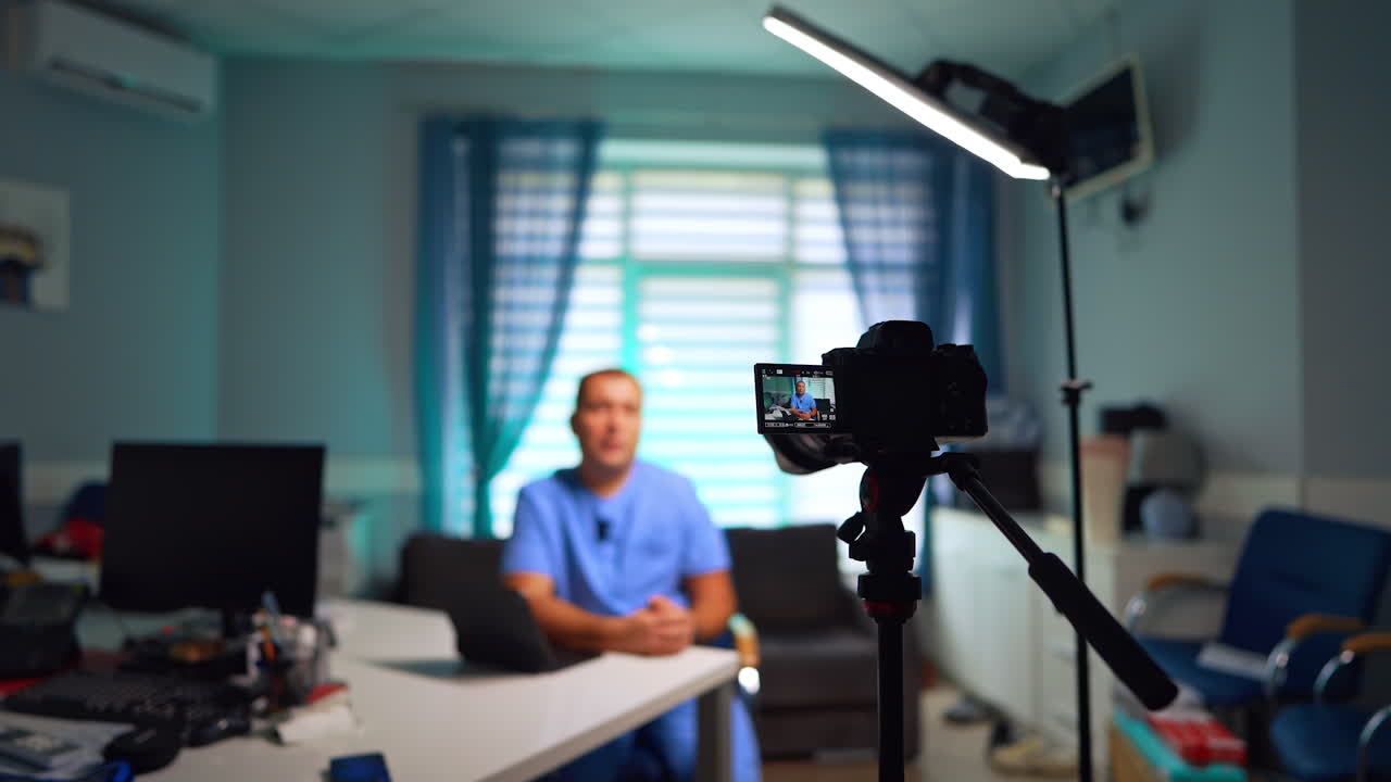 Camera and light set in the room directed on the man sitting at desk. Male doctor speaks to camera creating video content for vlog. Selective focus