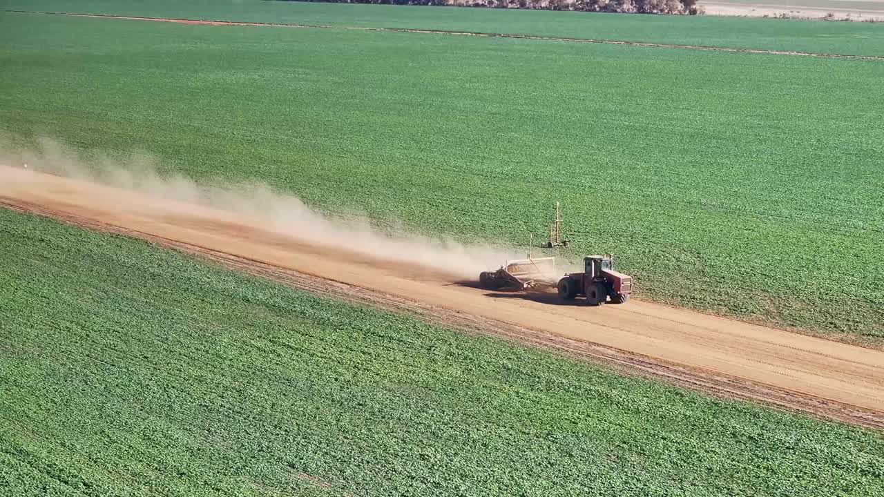 A tractor towing a small road grader along a dirt farm road between farm crops