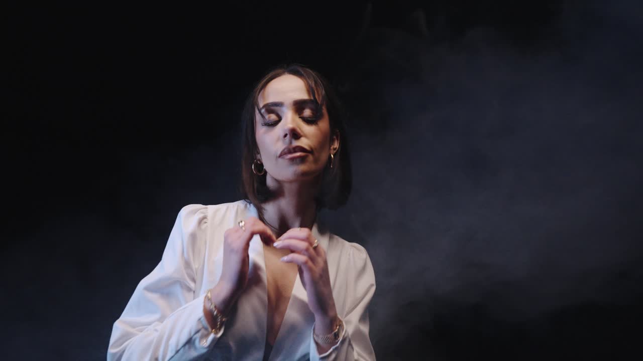 woman with a white outfit poses with a dark background. she tucks her hair behind her ears and looks serious