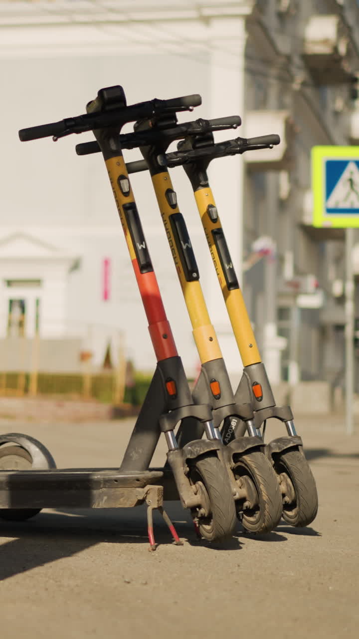 도심 횡단보도 근처 인도에 주차된 전기 스쿠터 행렬, 지나가는 흐릿한 보행자, 햇살이 비치는 아침 분위기, 줄지어 있는 렌탈 차량, 다가오는 배달 기사, 앱을 확인하는 통근자, 관광객