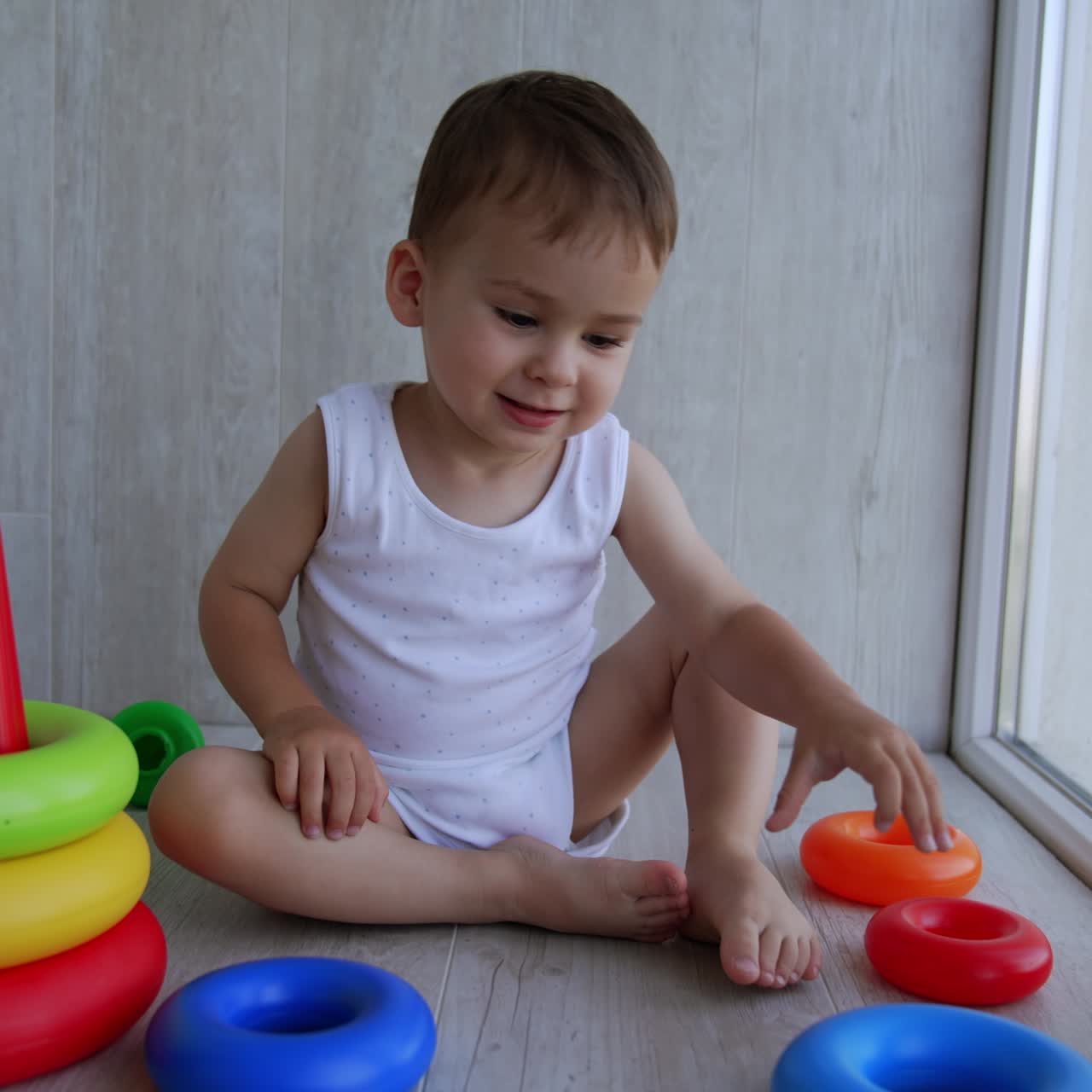 Active Caucasian toddler playing with his toys. Baby boy sits on the floor throwing the rings from toy pyramid