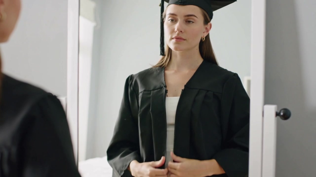 Donna in abito bianco aderente che si sistema mentre prova una toga nera davanti allo specchio, mettendo il tocco per la laurea.
