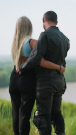 man driver with beloved girlfriend embraces near modern motorcycle standing on hill top grass against calm lake on spring day backside view slow motion
