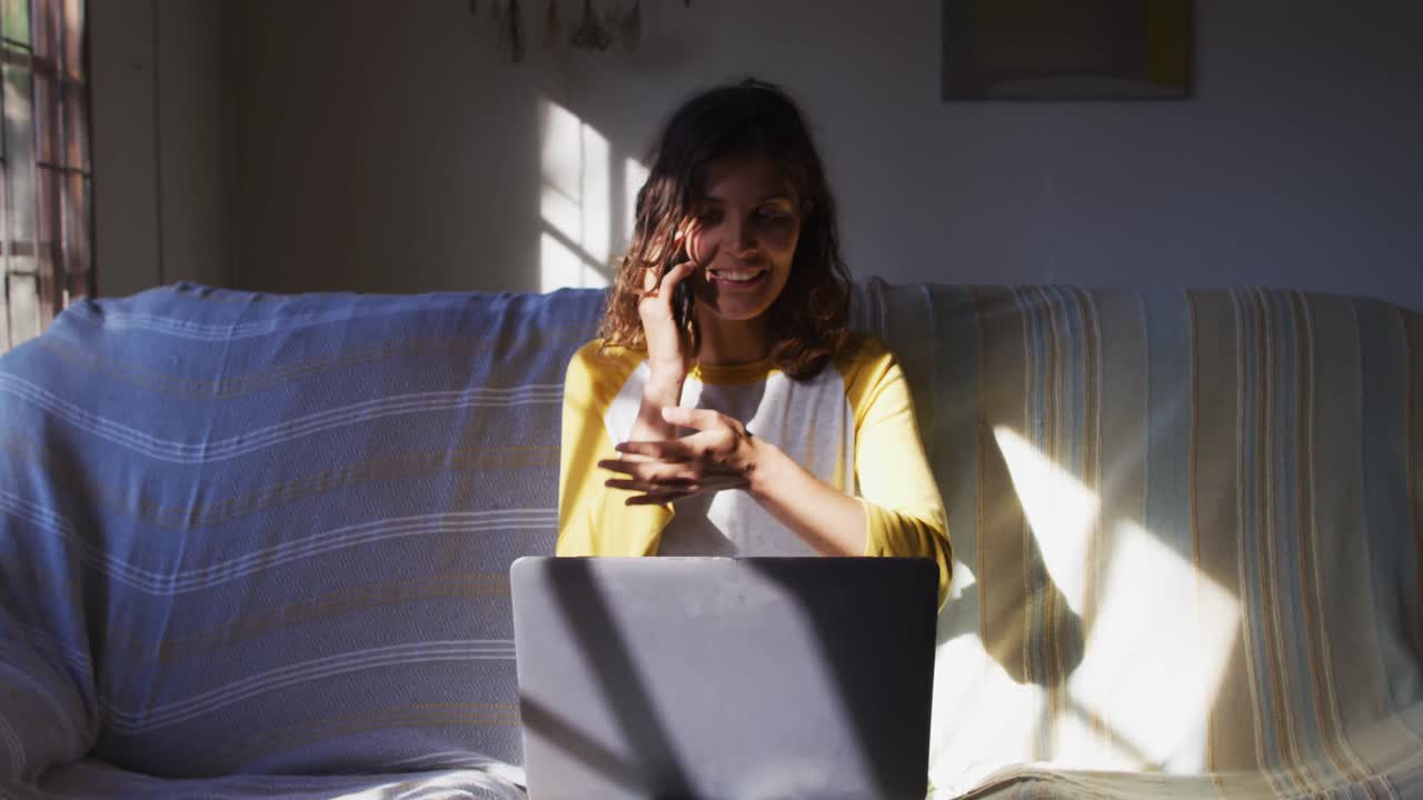 feliz mujer de raza mixta sentada en el sofá hablando en el teléfono inteligente usando una computadora portátil en la sala de estar soleada