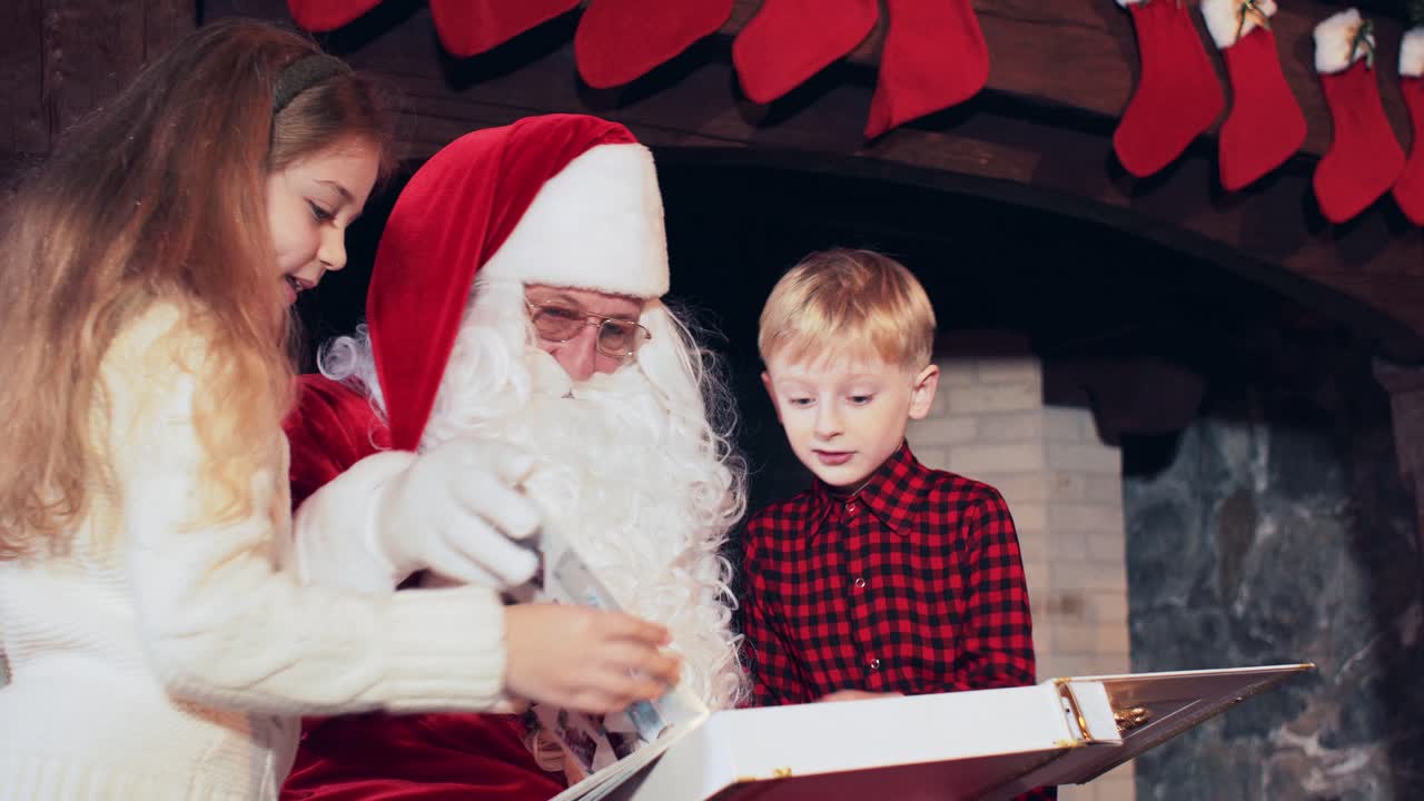 A boy and a girl are reading a book with Santa Claus in a room near the fireplace with Christmas stockings