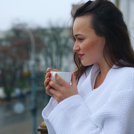 cheerful woman enjoying quality time in a hotel. young beautiful woman in bathrobe on balcony drinking coffee in the morning on the city background. side view