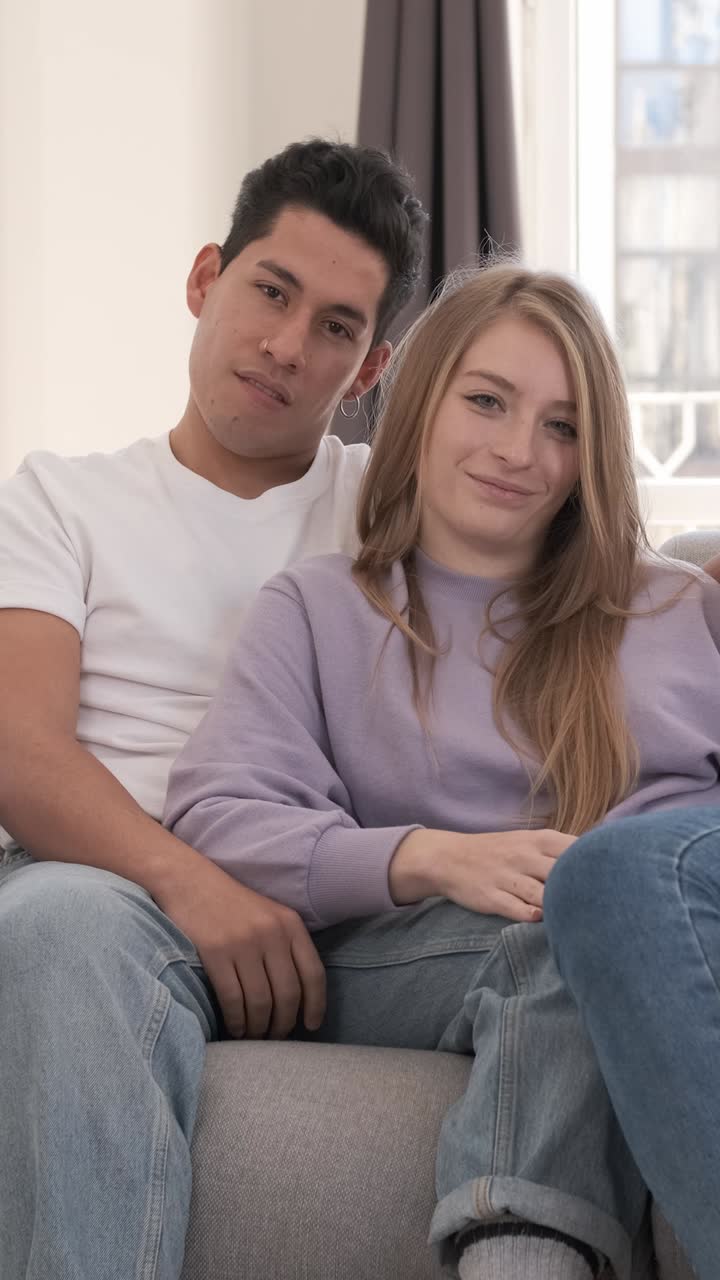 Ritratto di giovane coppia che sorride e guarda la fotocamera mentre è seduta sul divano di casa.