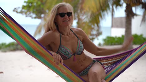 pretty mature woman in sunglasses and bikini in a hammock looking to the sky with arm up and then smiling at the camera in a portrait