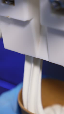 hand in blue gloves puts a paper cup under a food automat. soft creamy ice-cream is filling the cup close up. vertical video