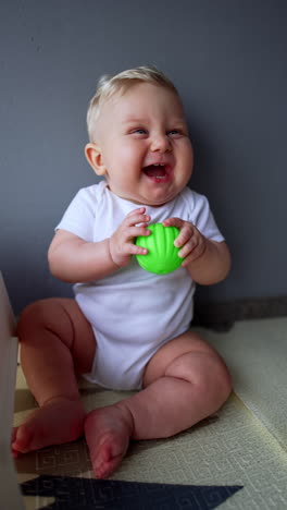 beautiful blond baby boy wearing white bodysuit sitting on the floor. kid is laughing adorably playing with a toy. vertical video.
