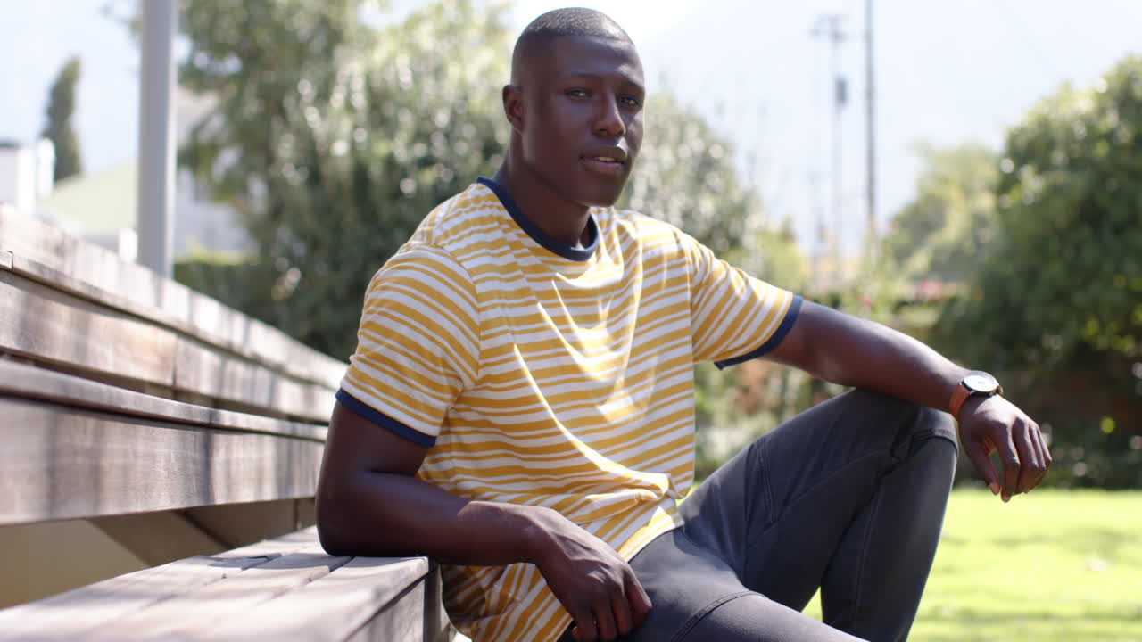 Relaxing outdoors, african american man sitting on bench in casual striped shirt, enjoying sunlight