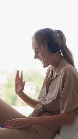 happy blonde woman in headphones communicates with friend during videocall via laptop waving hand against bright window at home slow motion
