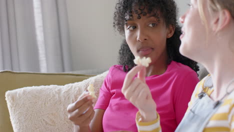 happy diverse teenage female friends talking and eating popcorn on couch at home, slow motion