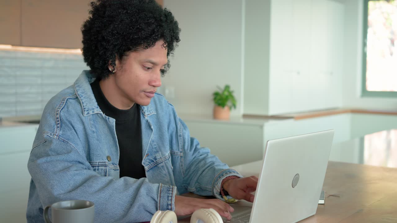 Mid adult man spotting laptop screen at kitchen island placing mug beside headphones typing notes