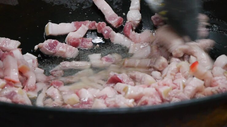 Potongan kulit babi mentah dan merah yang baru dimasukkan ke dalam wajan sedang ditumis dalam minyak panas mendidih, diaduk dengan spatula, direkam secara close-up.