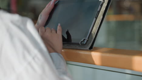 Close up back view of woman in white shirt operating digital tablet while leaning on wooden railing in brightly lit modern interior with natural light casting shadows on her hands and screen