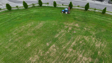 Aerial view revealing maintenance worker driving agricultural tractor, cutting roadside grass near Piacenza residential area under summer sunlight