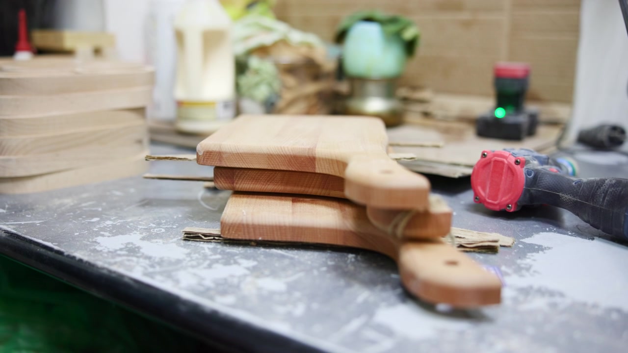 Taglieri di legno impilati su un banco da lavoro che mostrano una scena di bottega artigiana con polvere di levigatura, trapano rosso, sfocato.