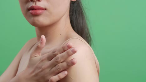 close up de una mujer asiática aplicando loción corporal a su hombro y sonriendo mientras está de pie en el fondo de la pantalla verde en el estudio