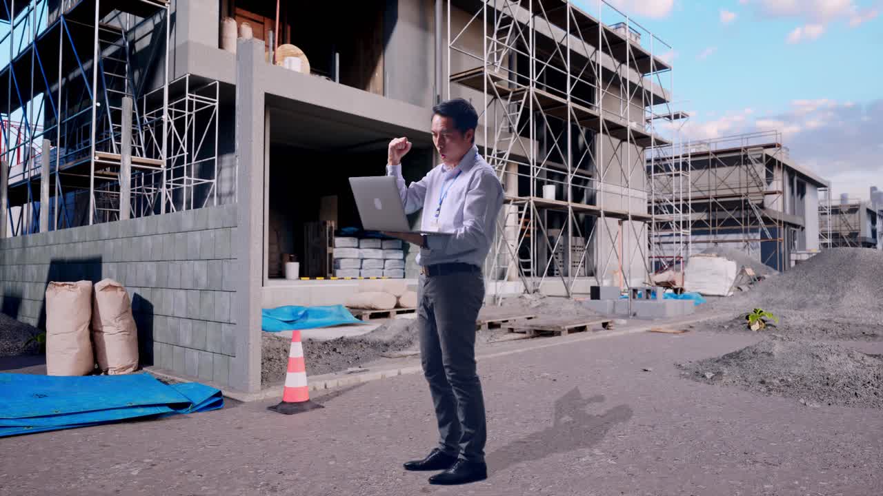 Full Body Side View Of An Asian Male Professional Worker Standing With His Laptop At Construction Site in Progress to New House, He Raises His Fist Up With Screaming Goal After Typed On The Laptop