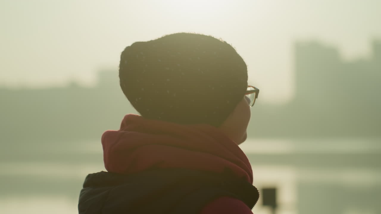 朝焼けの川辺に立つ、ビーニーをかぶったアジア人女性の横顔。眼鏡越しに霧がかったスカイラインを見つめ、柔らかな逆光が眼鏡に反射している。赤いスカーフとジャケットに身を包み、穏やかで物思いにふける様子。肌寒い都会の水辺の情景。