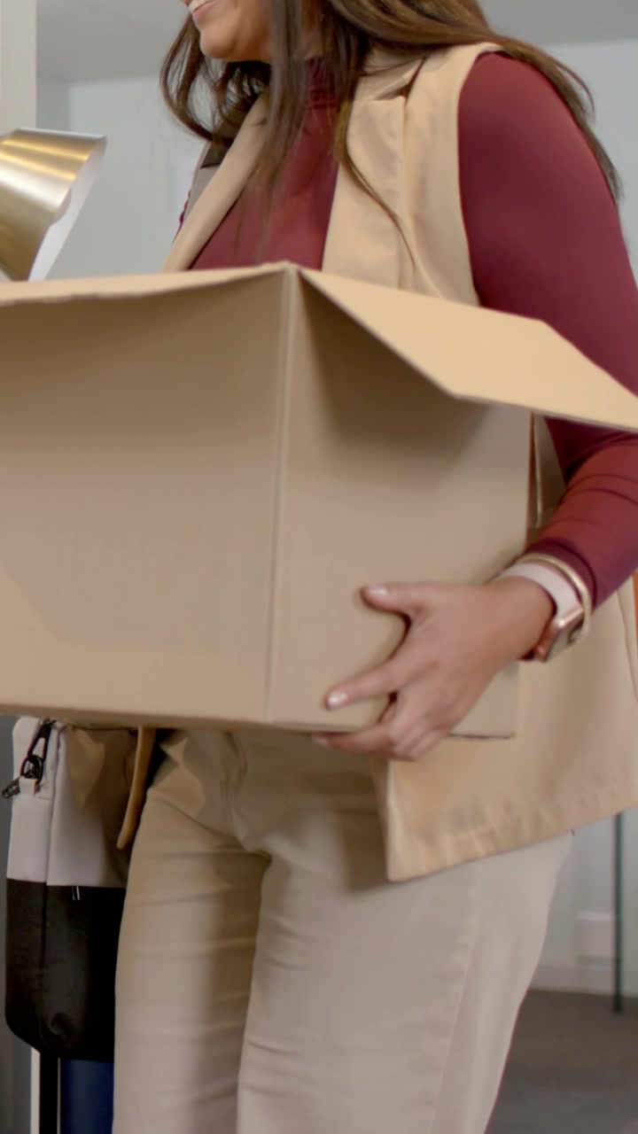 Video verticale: Donna con maglia bordeaux entra in ufficio, trasporta una scatola di cartone con una lampada di ottone che spunta.