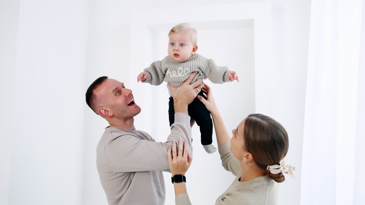 Caucasian parents are holding their lovely baby together. Father shakes his son and kid smiles adorably. White backdrop.