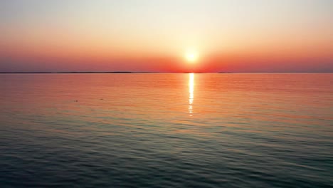 Left-Flying-Drone-View-of-Gorgeous-Ocean-Sunrise-with-Bright-Glowing-Sun-Casting-Colorful-Red,-Orange-Purple-and-Yellow-Reflections-Over-Peaceful-Rippling-Waves-of-the-Sea-with-Beautiful-Sky-and-Water