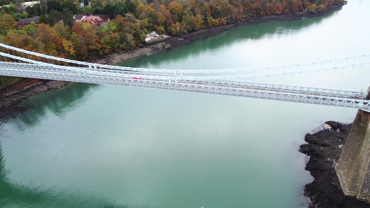 Aerial drone view of Menai Suspension Bridge with stone towers over water and second bridge in background showing engineering structure and coastal landscape